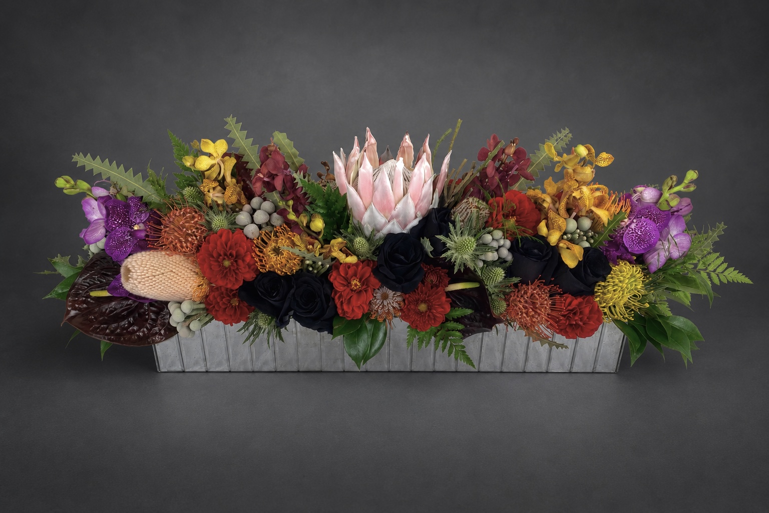 Long low arrangement of orchids, protea and dark roses in a rectangular gray vessel