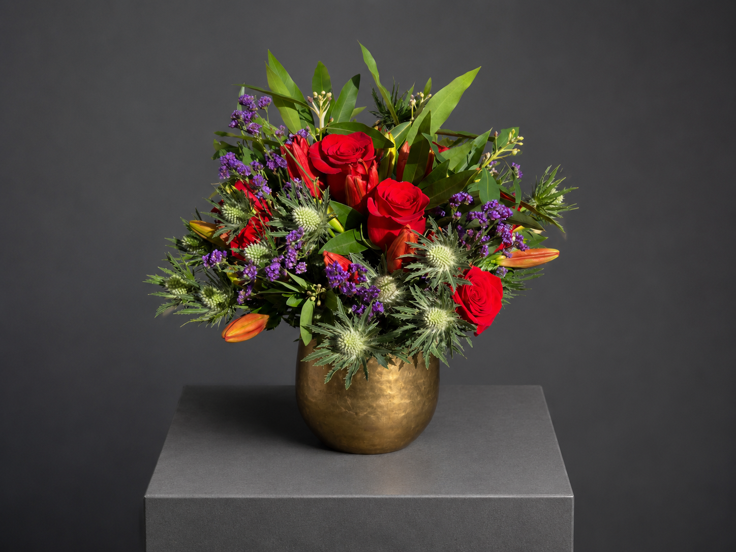 Red roses, spiky green flowers and purple accents in a gold-toned vase