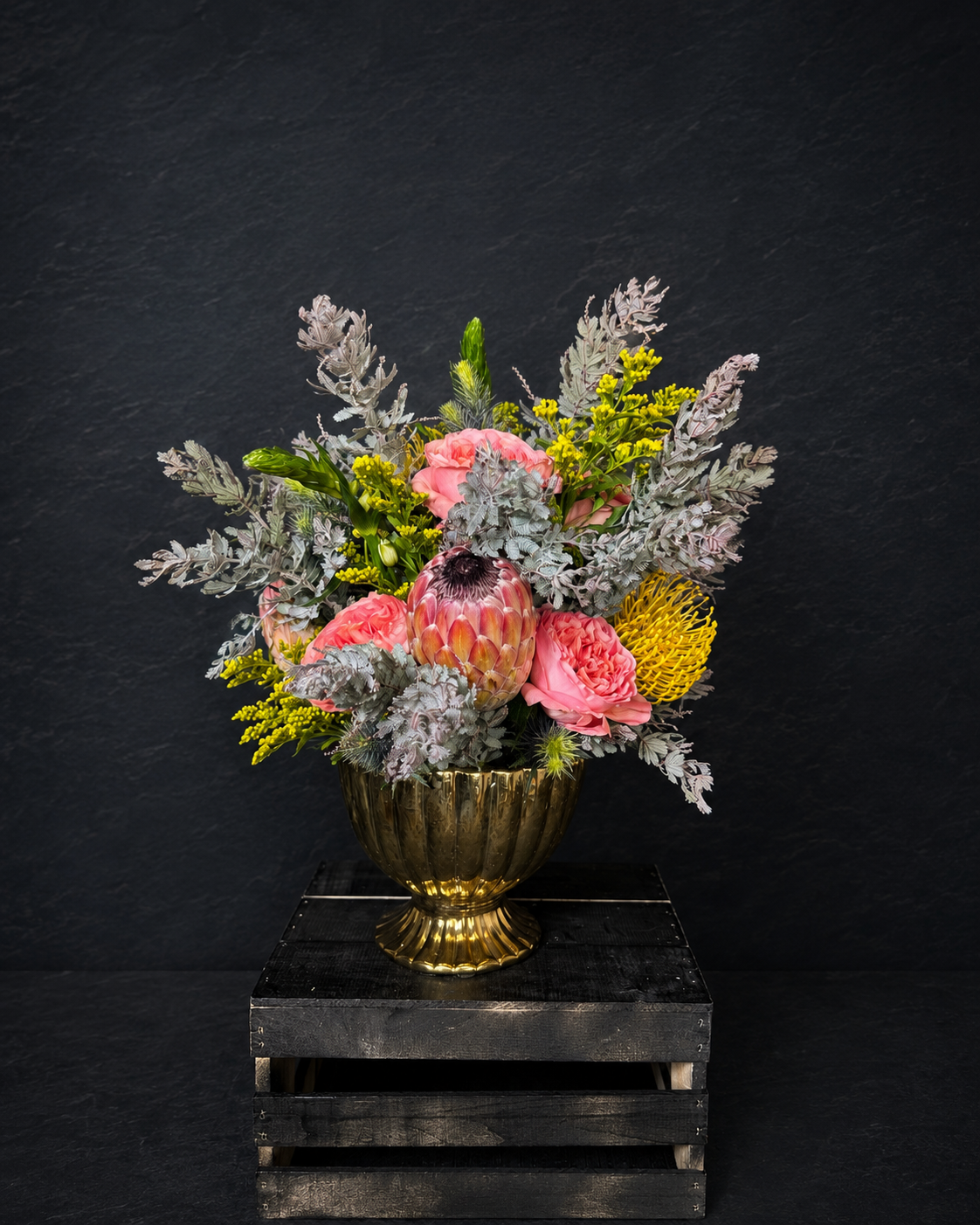 Pink roses and protea with silver foliage and yellow accents in a footed gold-toned vase