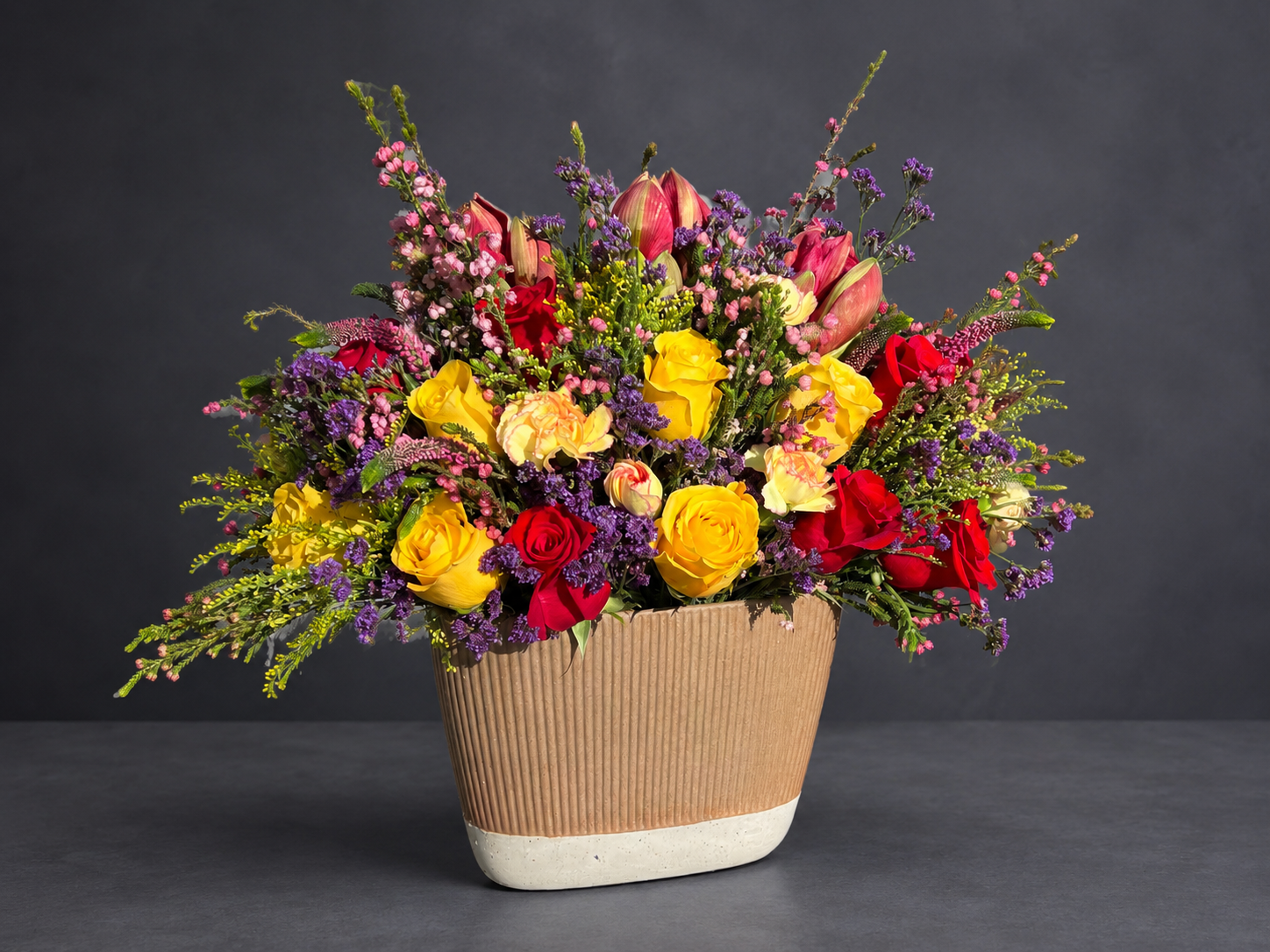 Red and yellow roses with pink and purple flowers in a ribbed ceramic vessel