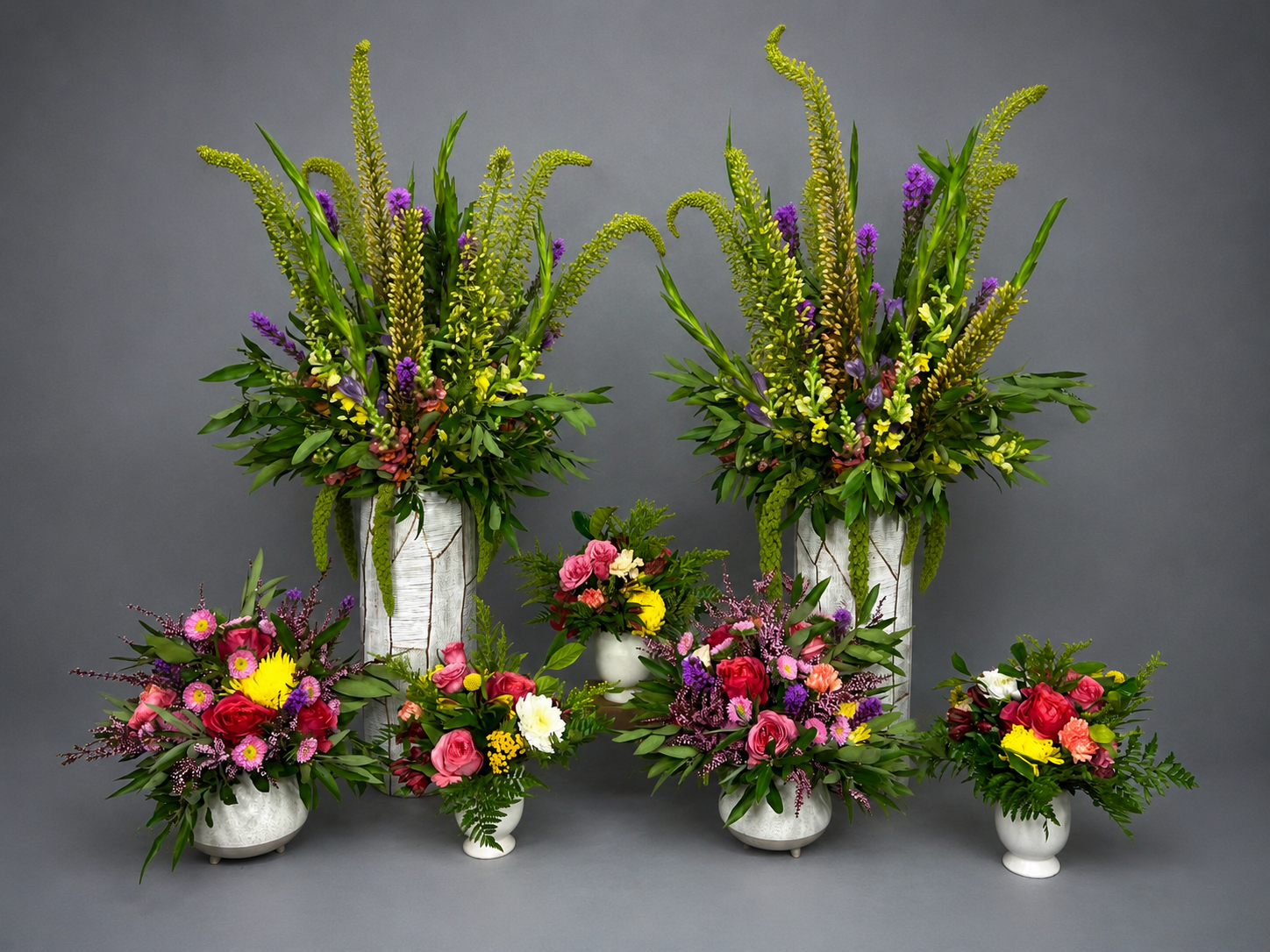 Two tall green and purple arrangements with five smaller colorful arrangements in pale vessels